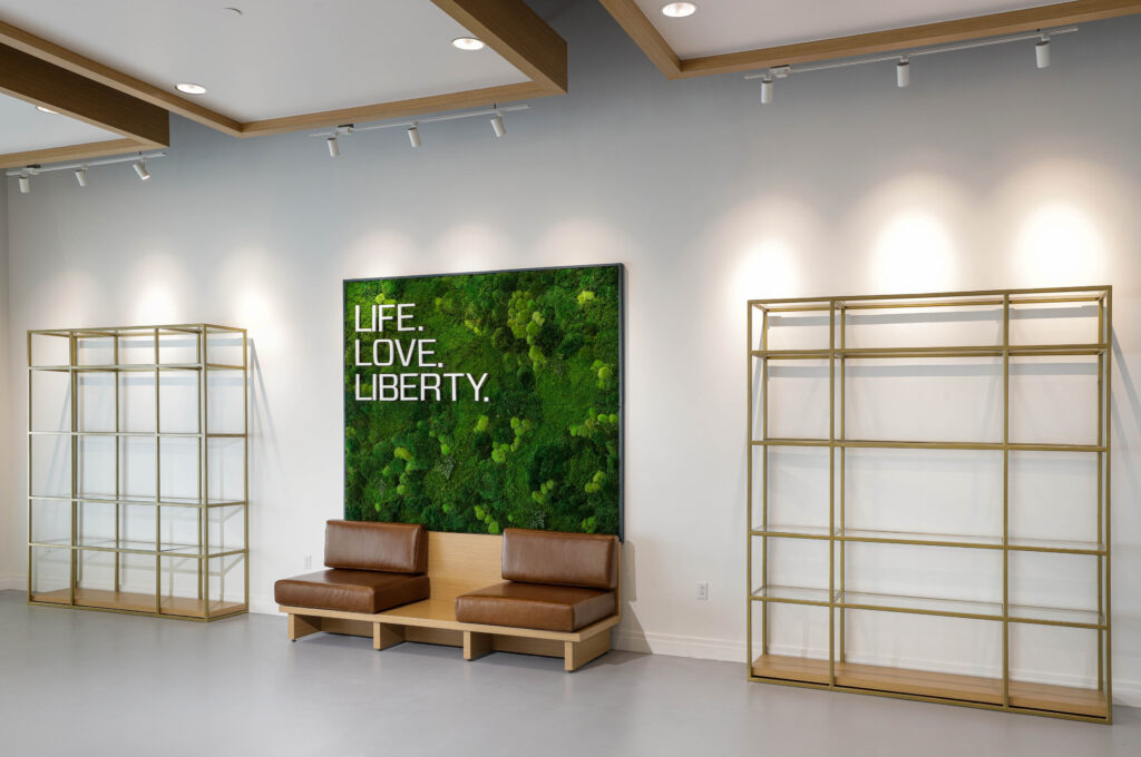 Two brown leather benches sit in front of a rectangular moss wall art piece that reads, “LIFE. LOVE. LIBERTY.” flanked by two empty metal display shelves.