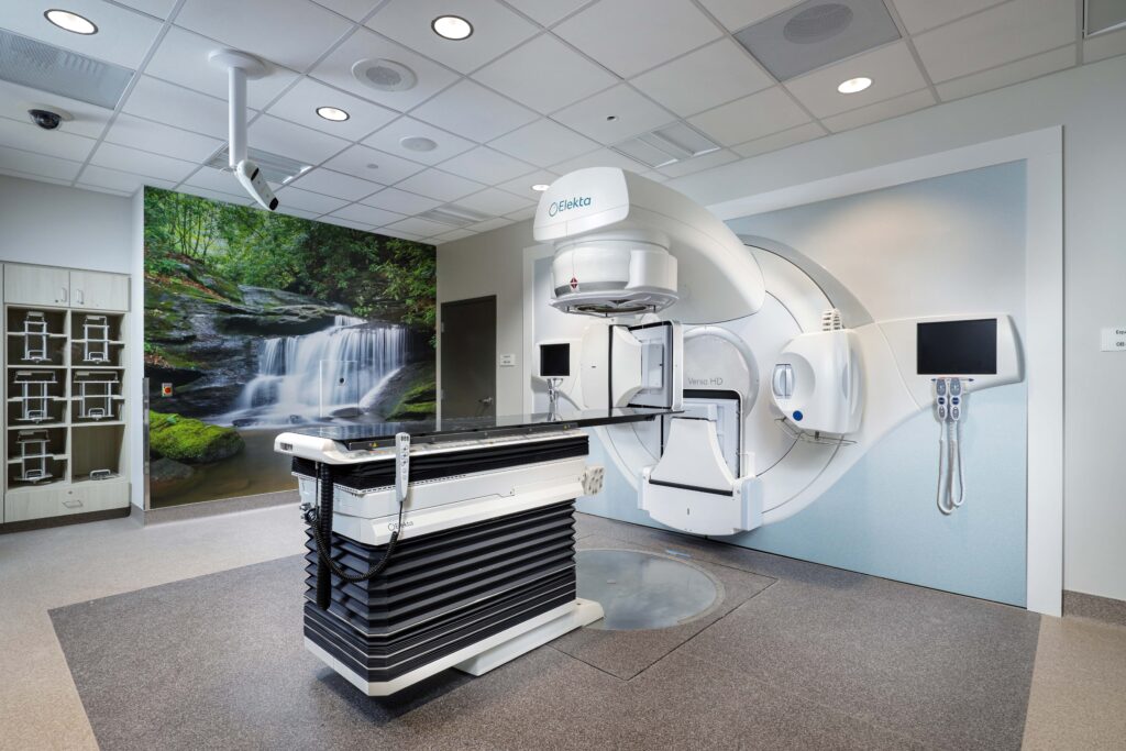 An Elekta Versa HD radiation therapy machine with an adjustable treatment table in a clinical room featuring a waterfall wall mural.