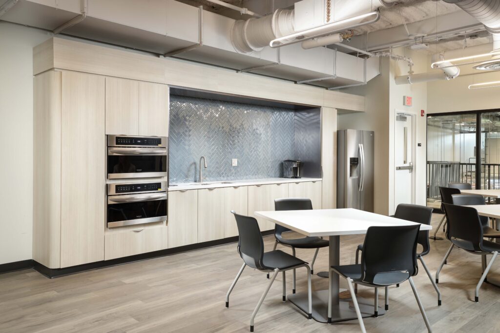 Modern office breakroom with light wood cabinets, stainless steel appliances, a gray herringbone tile backsplash, and a white square table with four black chairs.
