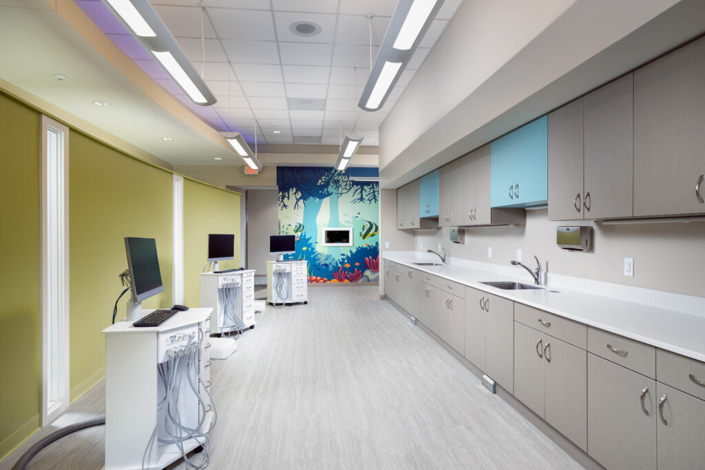 A dental treatment room with three computer stations and a long counter with sinks, featuring a wall mural of an underwater scene.