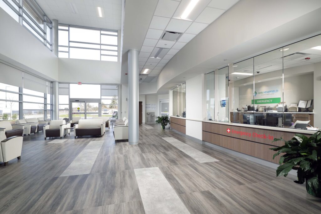 Modern hospital lobby with a waiting area and an Emergency Check-In desk behind a glass partition.