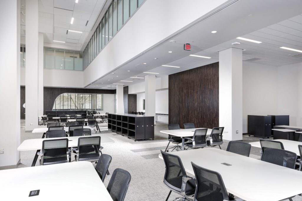 A modern, open-plan office space featuring rows of white desks with black rolling chairs, tall ceilings, and dark wood-paneled feature walls.