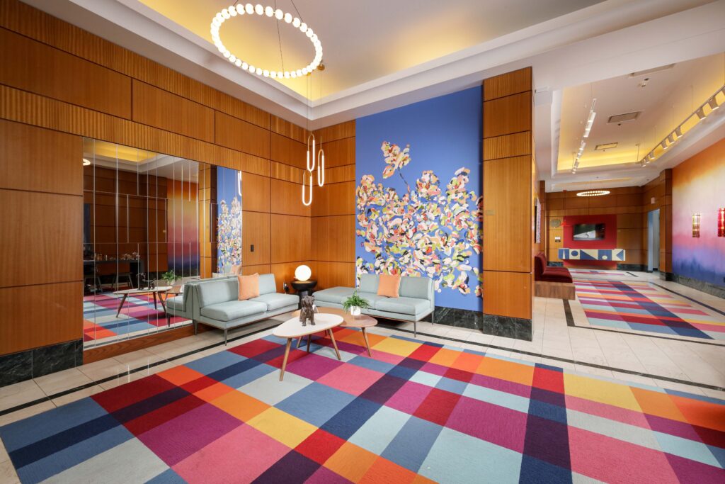 Modern hotel lobby with wood paneled walls, two light blue sofas, a colorful plaid carpet, and a large floral wall mural.