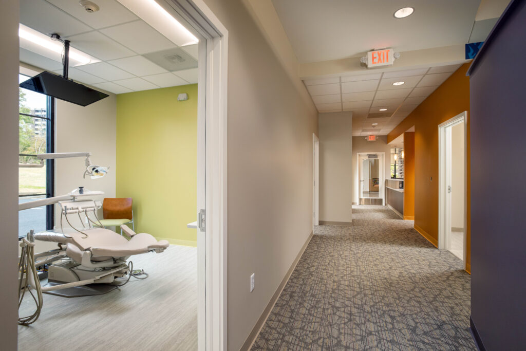 A dental exam room viewed from a hallway, with a dental chair and equipment visible through the open doorway.