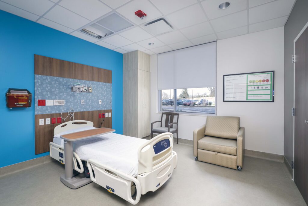 Hospital room with an adjustable bed, overbed table, a recliner, a side chair, and a window overlooking a parking lot.