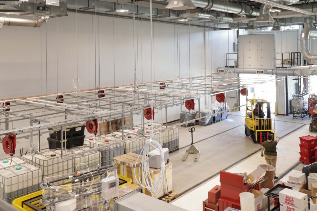 A warehouse interior with a yellow forklift being operated near rows of industrial storage containers and overhead red hose reels.