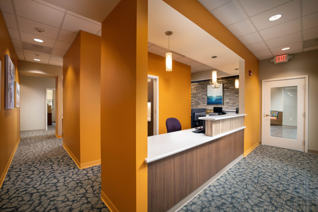 Reception desk with a white counter and wood-paneled front, set against a bright orange wall in an office lobby with patterned carpet.