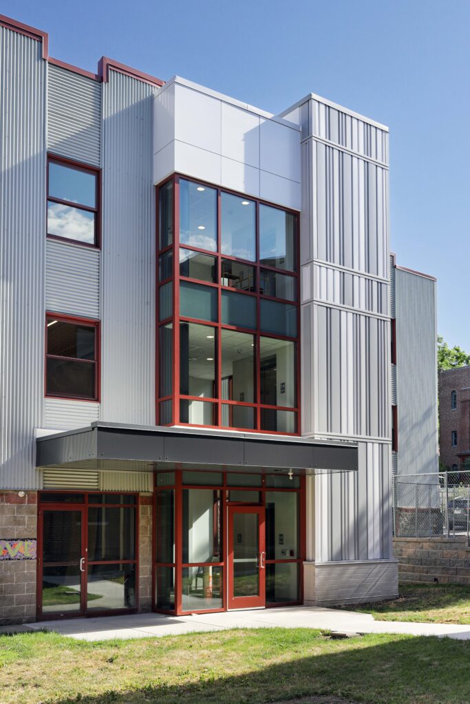 Modern three-story building exterior with corrugated metal siding, red window frames, and a glass-enclosed entrance.