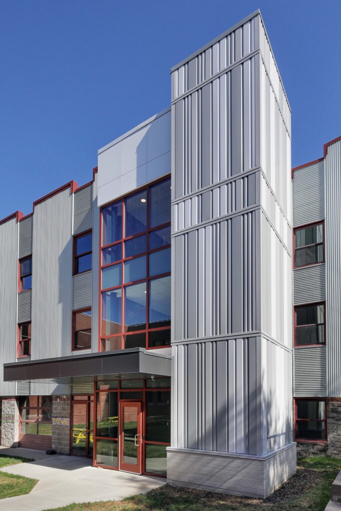 A modern three-story building exterior with vertical light gray siding, a central glass entrance tower, and red window frames.