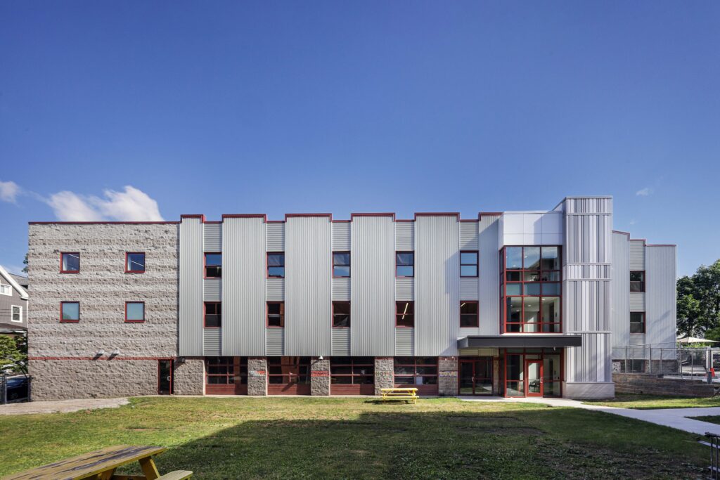 Modern three-story building with stone and metal siding, featuring a glass-enclosed stairwell and a yellow picnic table on the front lawn.