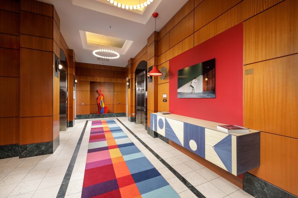 A modern hallway with wood-paneled walls, a red accent wall featuring art, a colorful geometric runner rug, and a console table.
