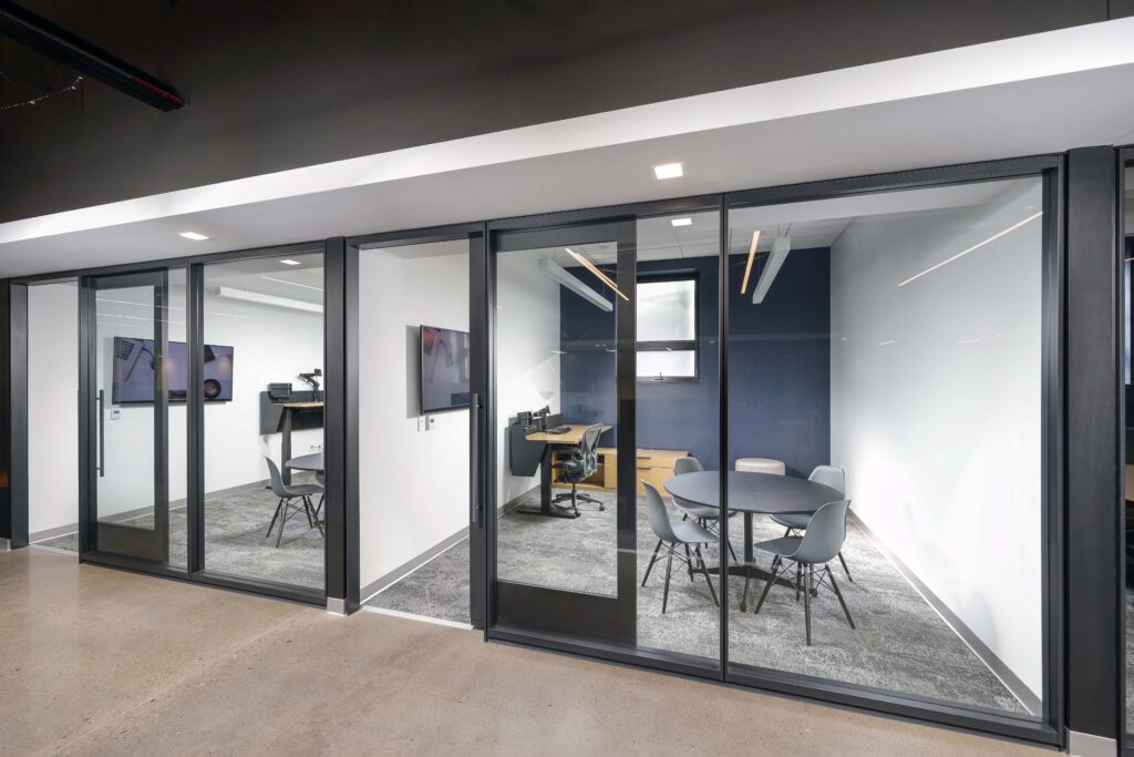 Two glass-walled office meeting rooms featuring small round tables, chairs, and individual desks.