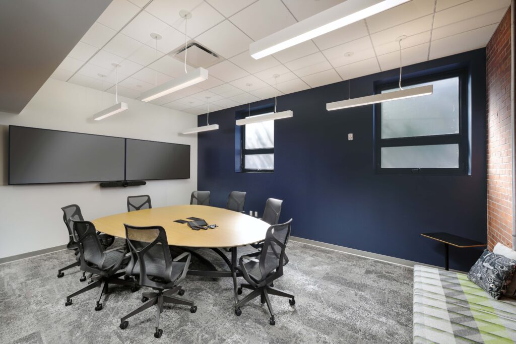 A modern conference room with a large oval table, six mesh office chairs, and two wall-mounted flat-screen monitors.