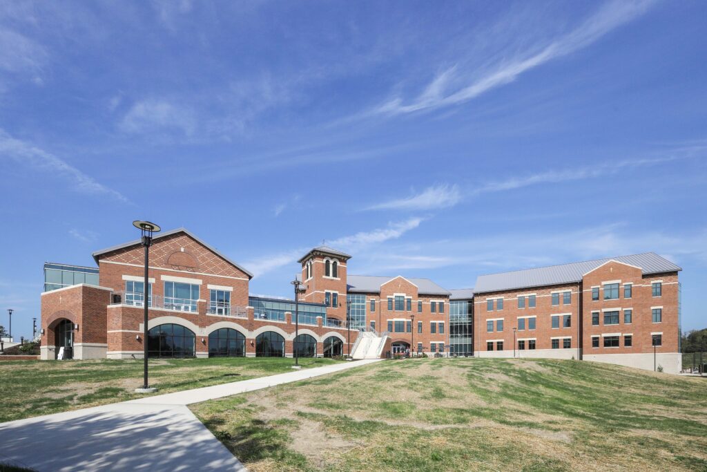A multi-story brick institutional building with a central tower and a paved path leading through a grassy lawn under a blue sky.