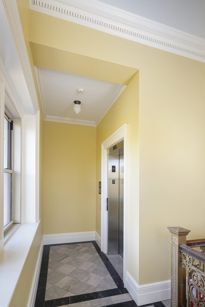 Elevator with a stainless steel door in a yellow-walled hallway with a tiled floor.