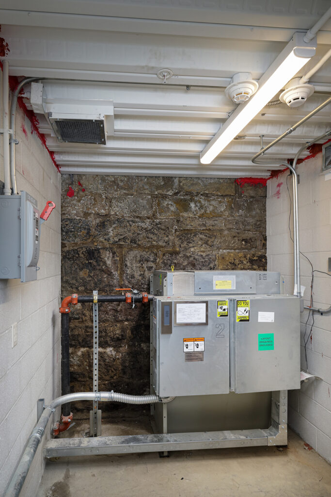A large industrial metal cabinet sits against a stone wall, connected to a black pipe with a red valve.