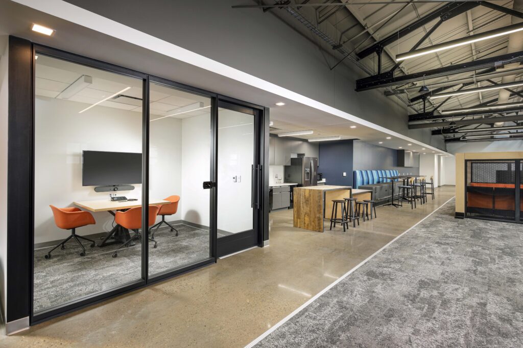 A glass-enclosed meeting room with orange chairs and a wall-mounted monitor, next to an open office kitchenette with high-top seating.