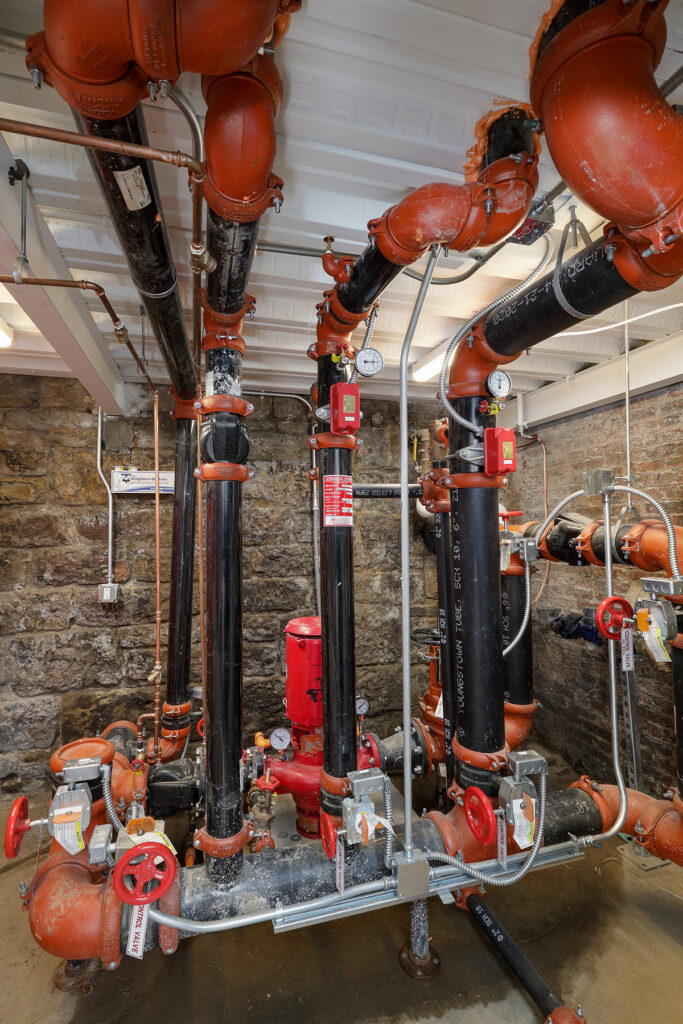 Industrial fire sprinkler piping system with red valves and gauges mounted against a stone wall.
