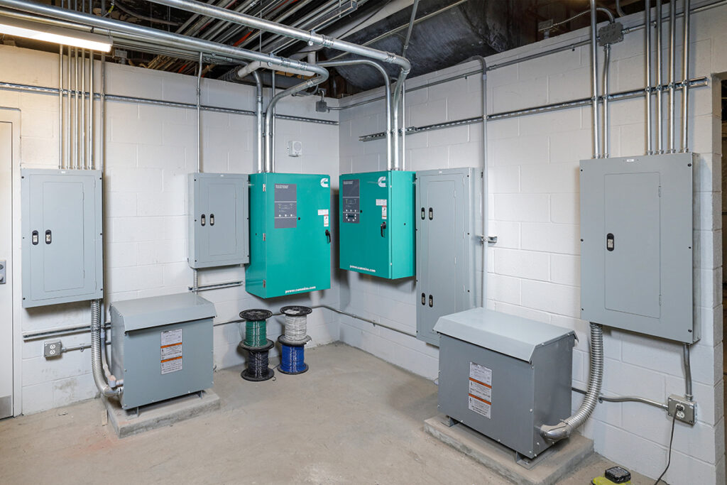 Electrical room with multiple grey cabinets, two green Cummins control units on the wall, and spools of electrical wire on the floor.