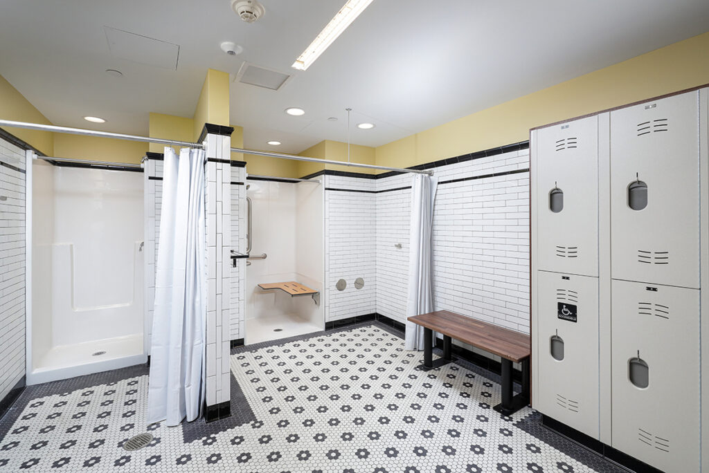 A locker room with two shower stalls, a wooden bench, and a bank of metal lockers on a patterned tile floor.