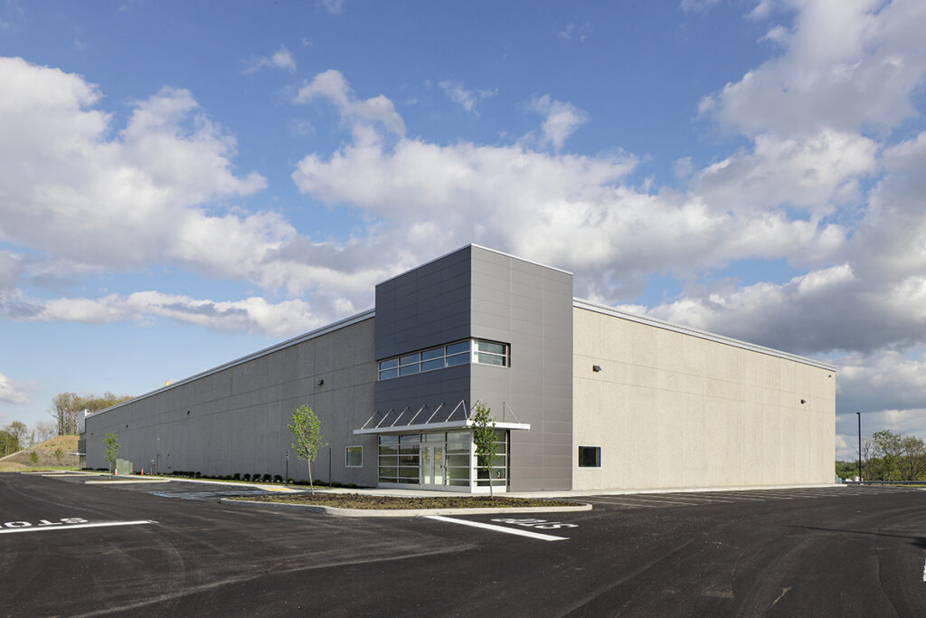 Modern commercial warehouse building with a gray facade and a glass entrance, set in an empty asphalt parking lot under a blue sky.