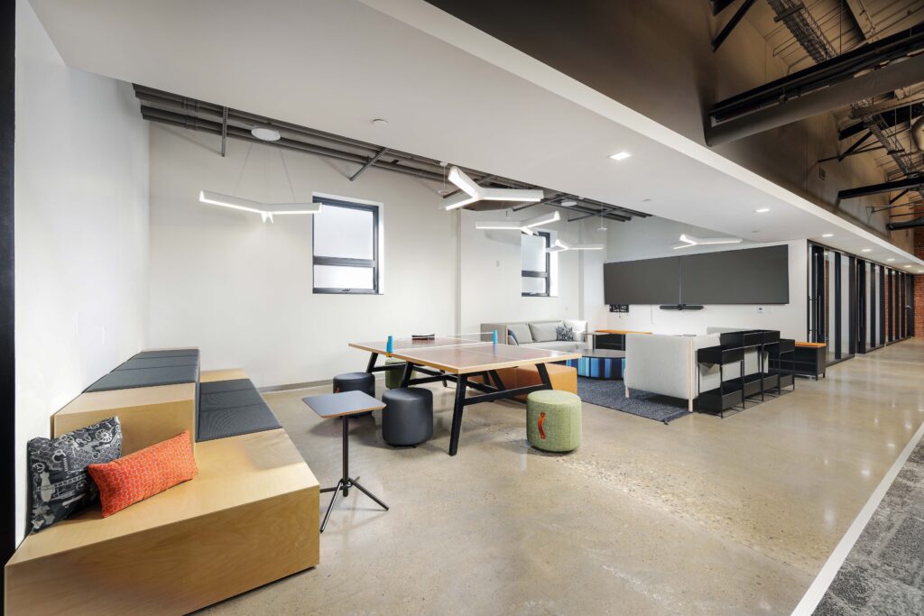 Modern office break room with a ping pong table, tiered bench seating, and various stools and sofas.