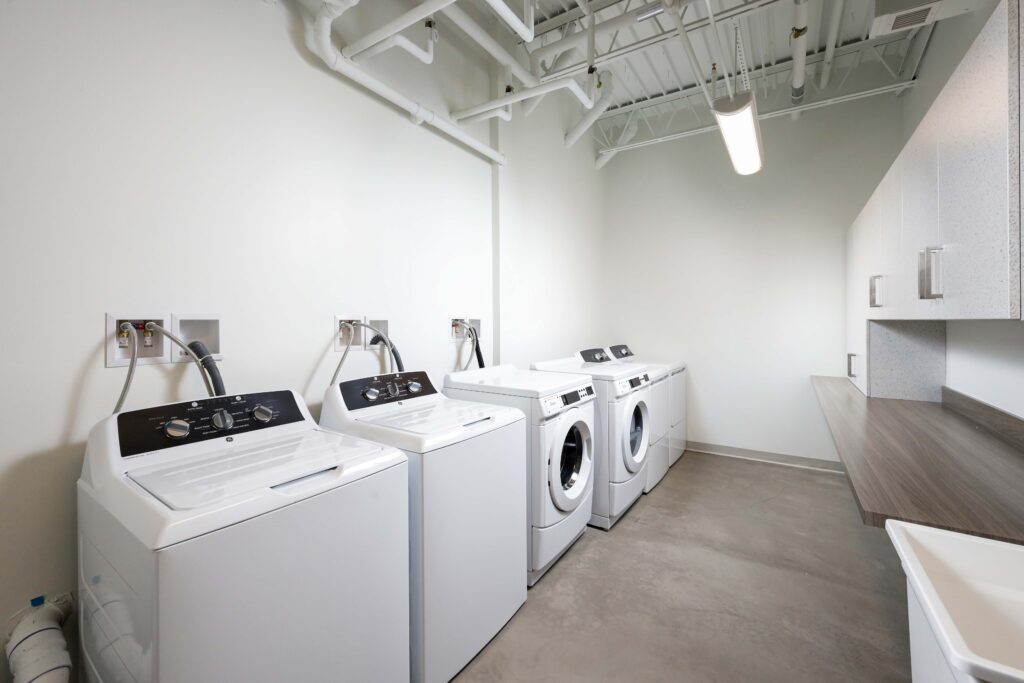 A laundry room with four white washing machines lined up against a wall, next to a counter and sink.