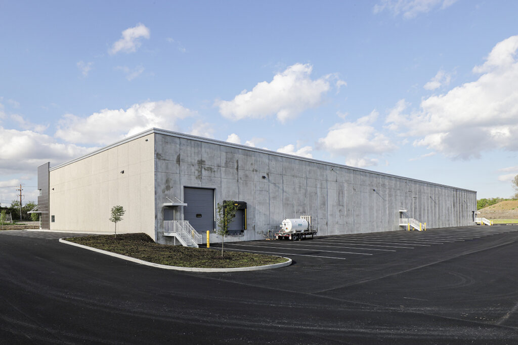 A large concrete warehouse building with multiple loading docks and an adjacent asphalt parking lot under a blue sky with scattered clouds.