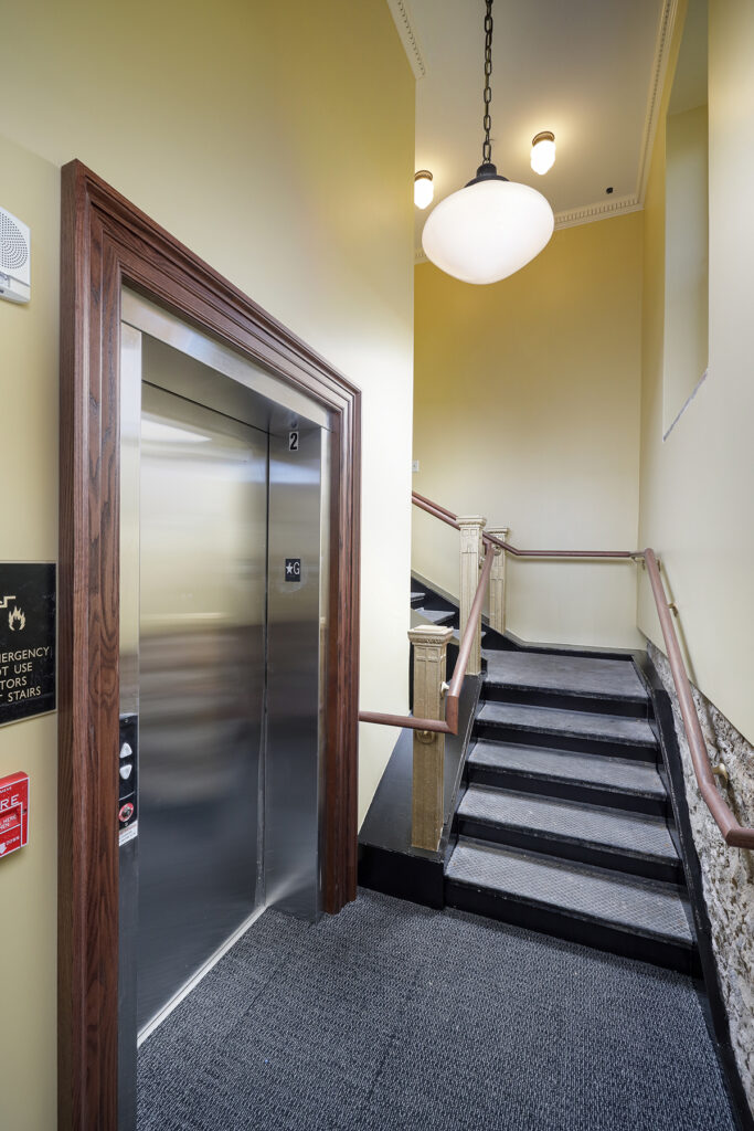 A stainless steel elevator door beside a staircase with a wooden handrail in a yellow-walled hallway.