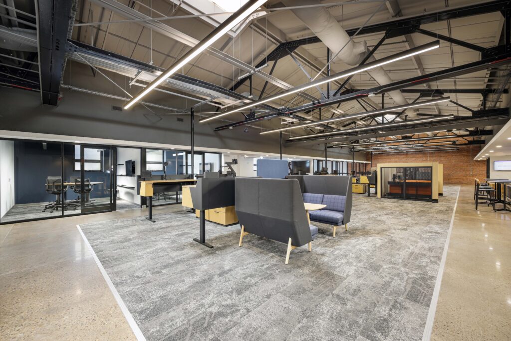Open-plan office with high ceilings, grey carpeted workstations, a booth-style seating area, and private glass-walled offices.
