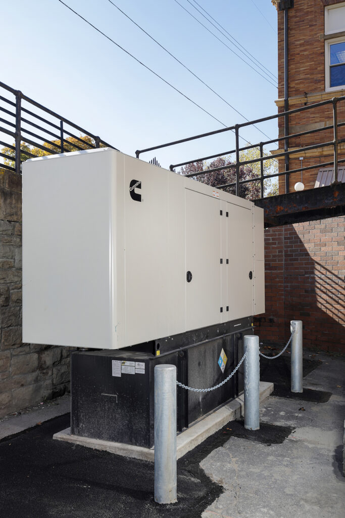 Cummins industrial generator in a white enclosure, installed on a concrete pad with a chain barrier.