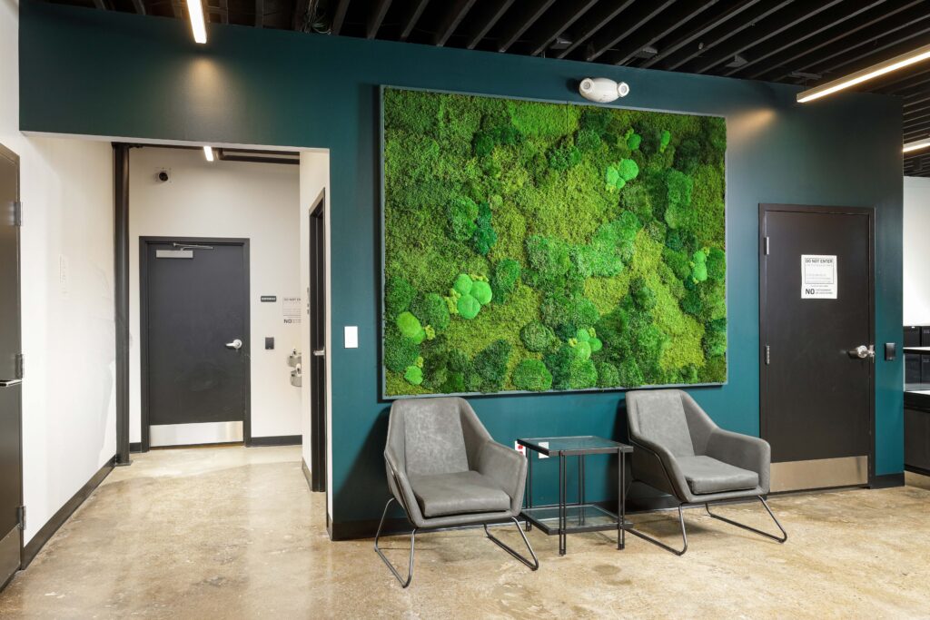 Two gray chairs and a small side table placed in front of a large, rectangular moss wall installation on a dark green wall.