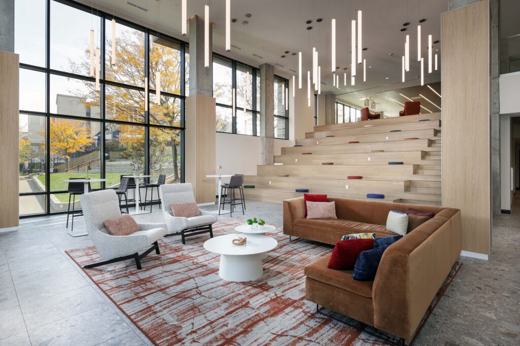 A modern lounge area with tiered wooden seating, a brown sectional sofa, two armchairs, and floor-to-ceiling windows overlooking a park.