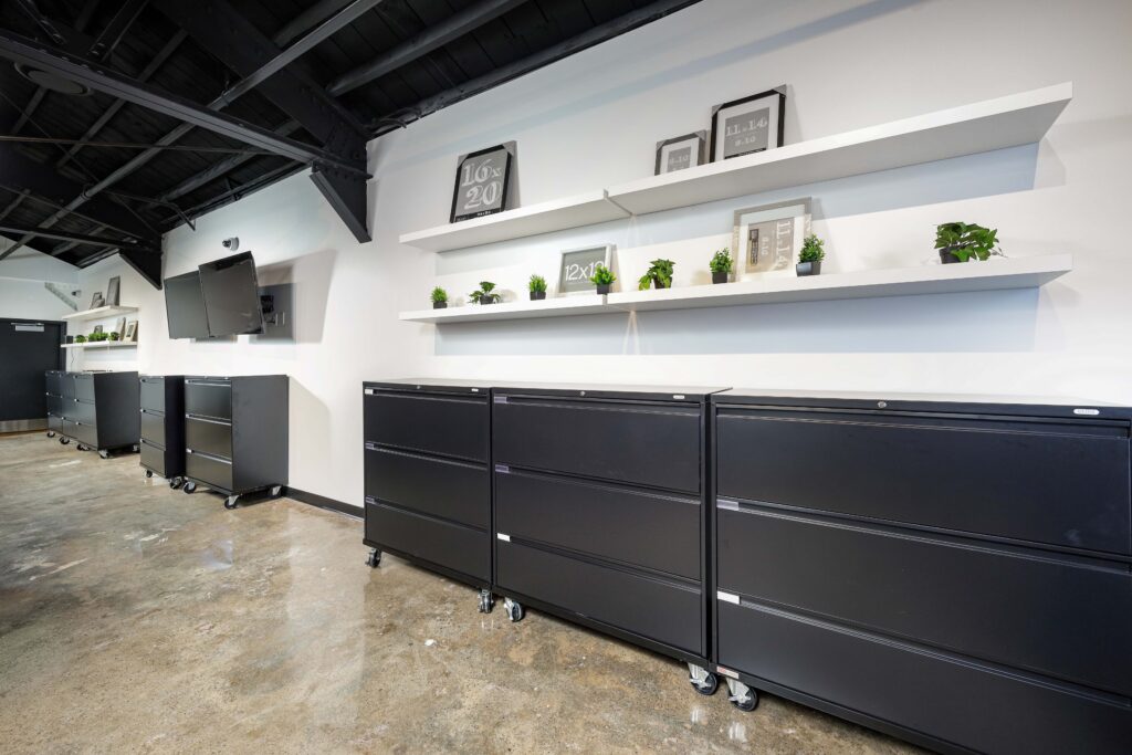 Black rolling filing cabinets stand along a white wall beneath two shelves displaying various picture frames and small potted plants.