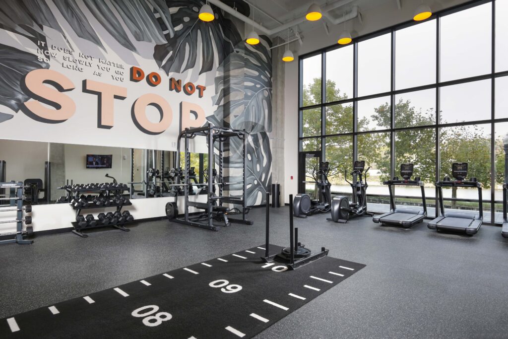 Gym interior with weights, a squat rack, elliptical machines, treadmills, and a marked track on the floor, featuring a large mural on the wall that reads “Do not stop.”