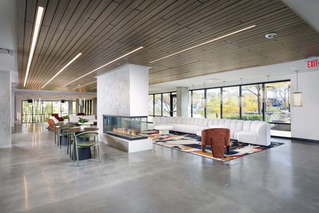 A modern lounge area with a central glass fireplace, white sectional sofa, round tables with green chairs, and large windows.