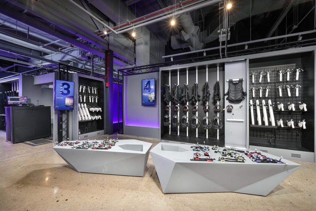 A virtual reality arcade room featuring tables covered in controllers and wall-mounted racks displaying VR headsets and haptic vests.