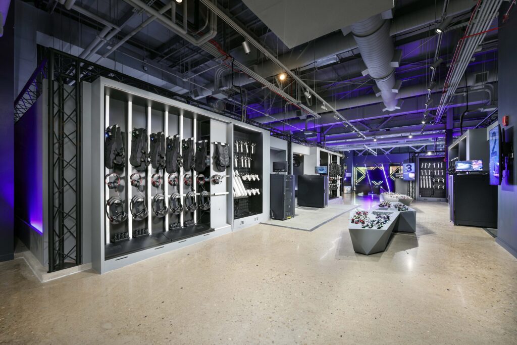 A VR arcade facility interior featuring wall-mounted headsets and vests, with controllers displayed on a central pedestal.