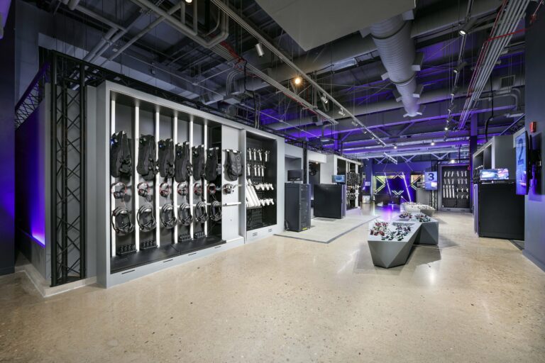 A VR arcade facility interior featuring wall-mounted headsets and vests, with controllers displayed on a central pedestal.