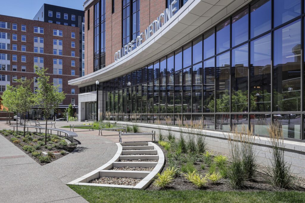 Exterior of the College of Medicine building, featuring a glass facade, curved concrete steps, and landscaped garden beds.