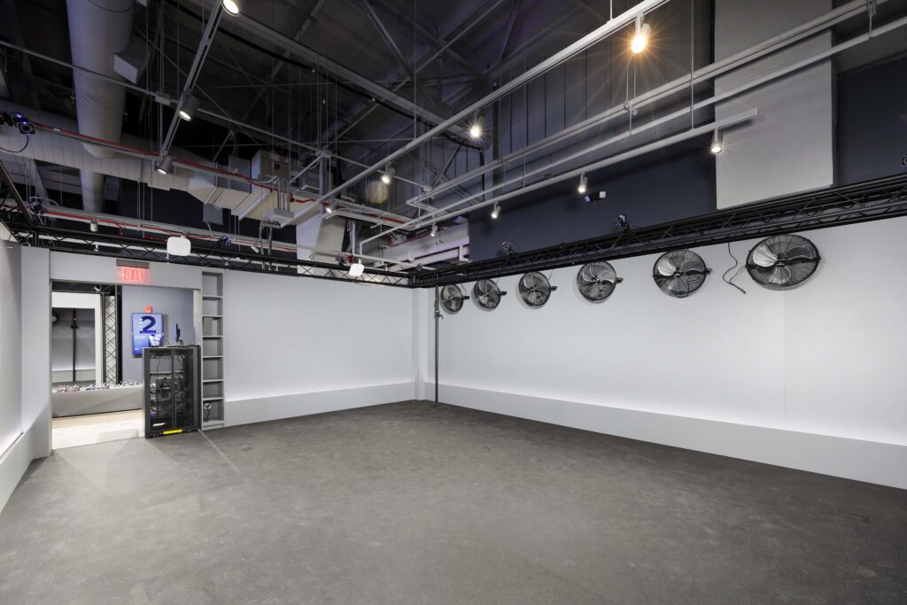 An empty studio space with white walls, a dark floor, and five large fans mounted on the wall under a metal lighting truss.
