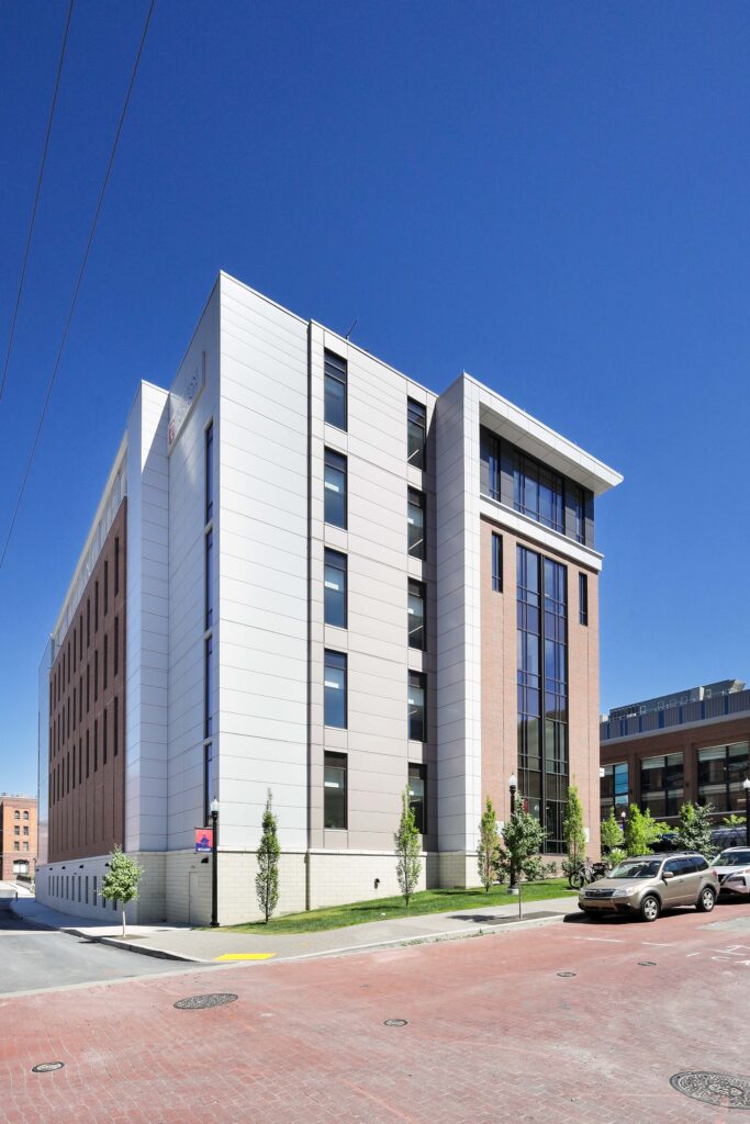 A modern, multi-story building featuring white panels and brick sections, located on a brick-paved street under a clear blue sky.