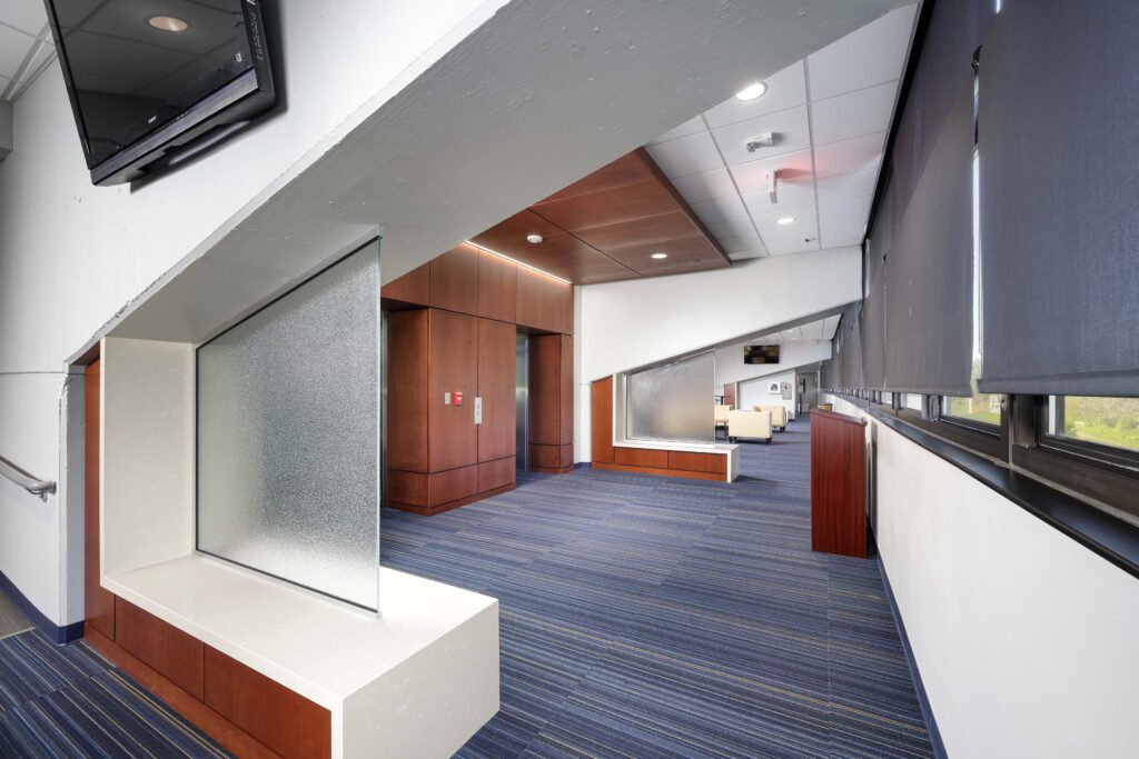 A modern office hallway with blue-striped carpeting, wood paneling, and glass dividers.