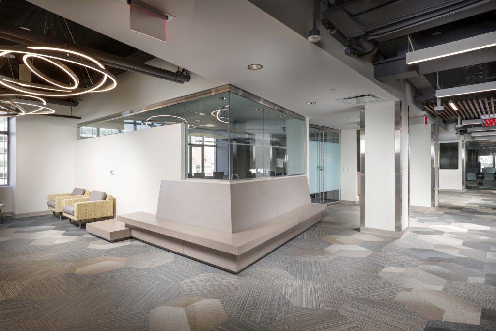 Modern office interior featuring a central glass-enclosed meeting room, two lounge chairs, and geometric carpet flooring.