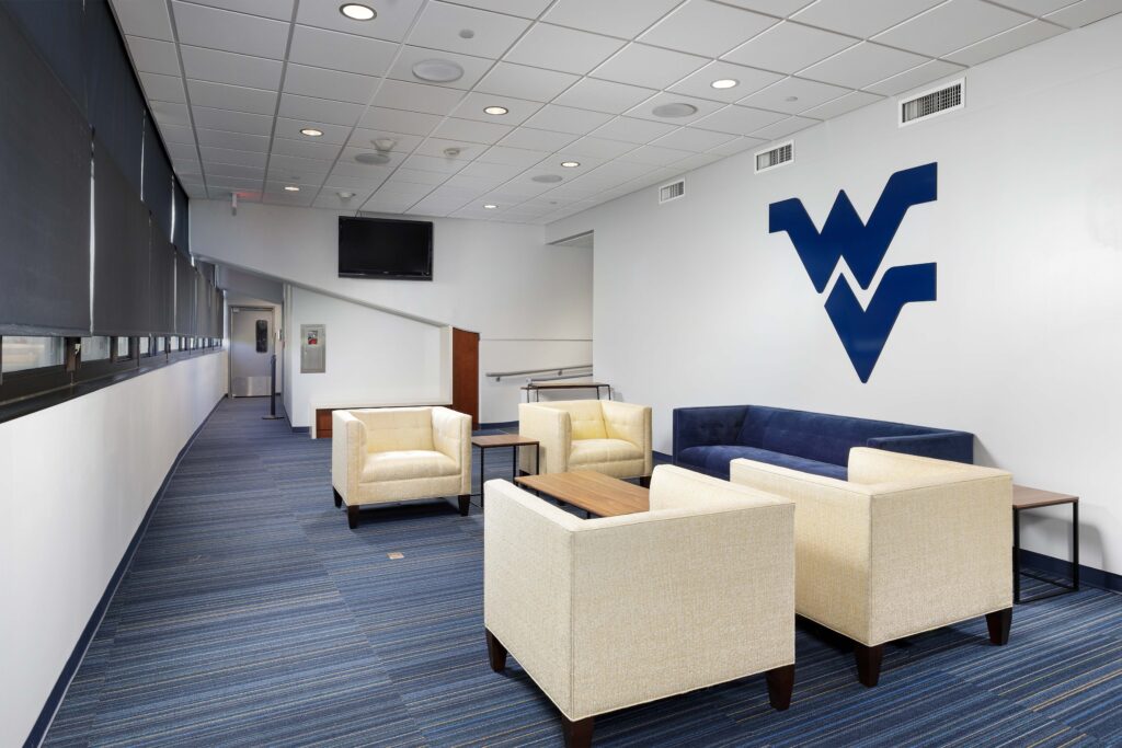 Lobby seating area with four light-colored armchairs, a blue sofa, and a large West Virginia University logo on the wall.