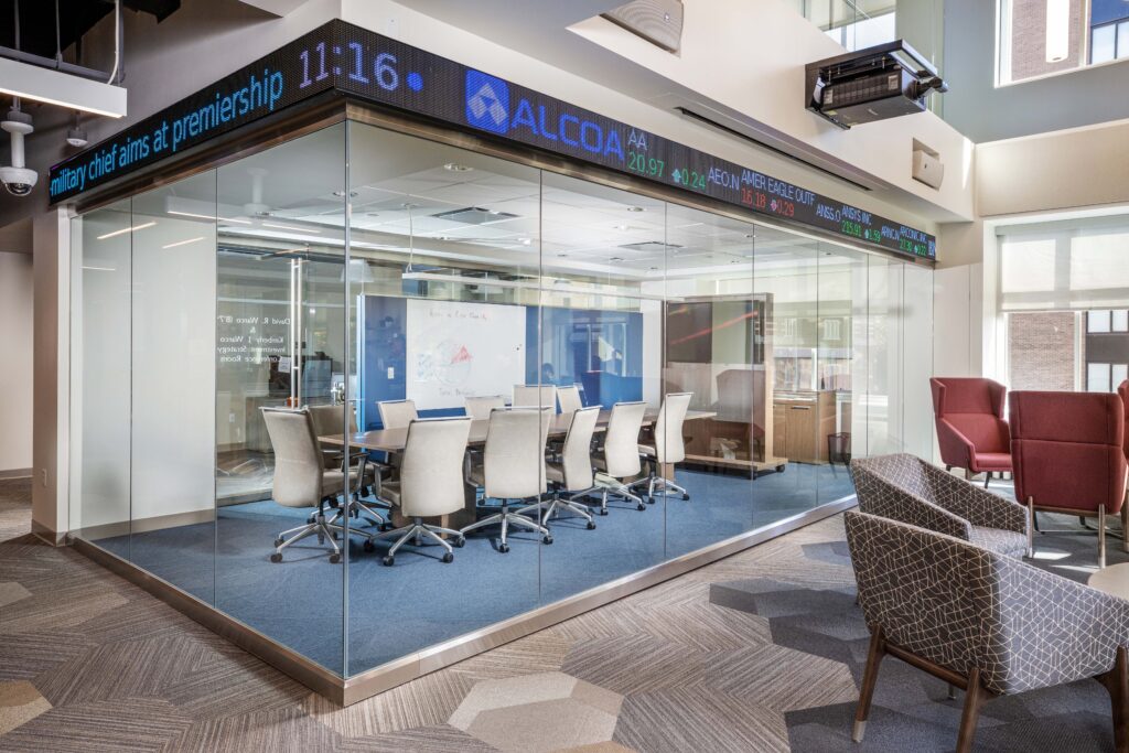A modern glass-walled conference room with a table and chairs, featuring a digital stock ticker running along the top of the walls.