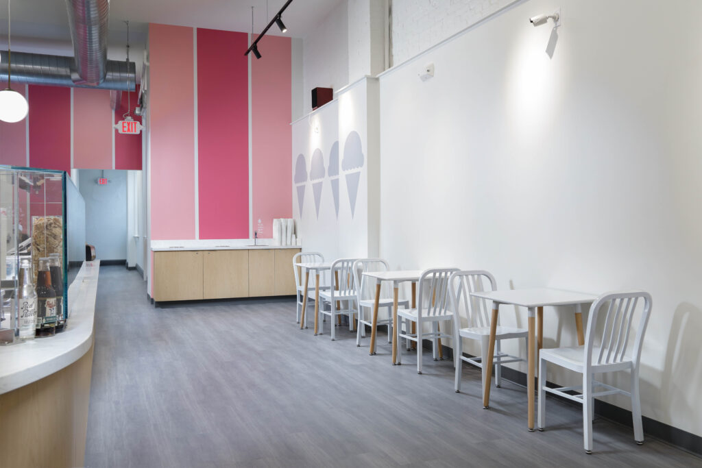 Interior of an ice cream shop with a pink accent wall, ice cream cone decals, and rows of white tables and chairs.