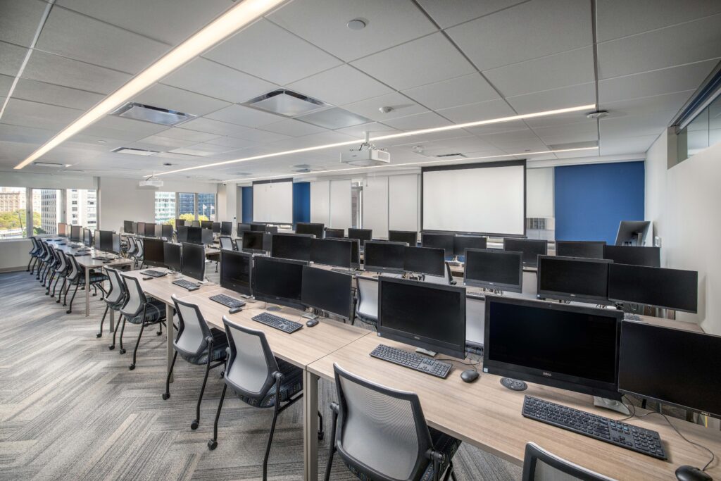 A computer lab with rows of desks, each equipped with a monitor, keyboard, and rolling chair, facing two projection screens at the front of the room.