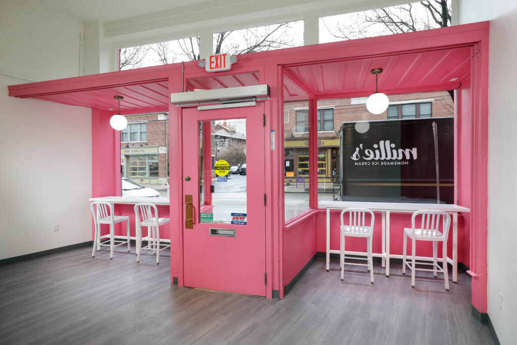 Interior view of a shop with a bright pink door and matching window framing, featuring white bar stools at counters and a view of a Millie’s Ice Cream truck through the glass.