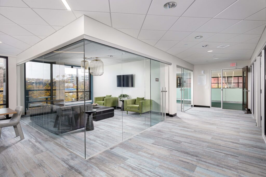 A glass-walled office suite furnished with a dark sofa and two green armchairs, situated in a modern commercial hallway.